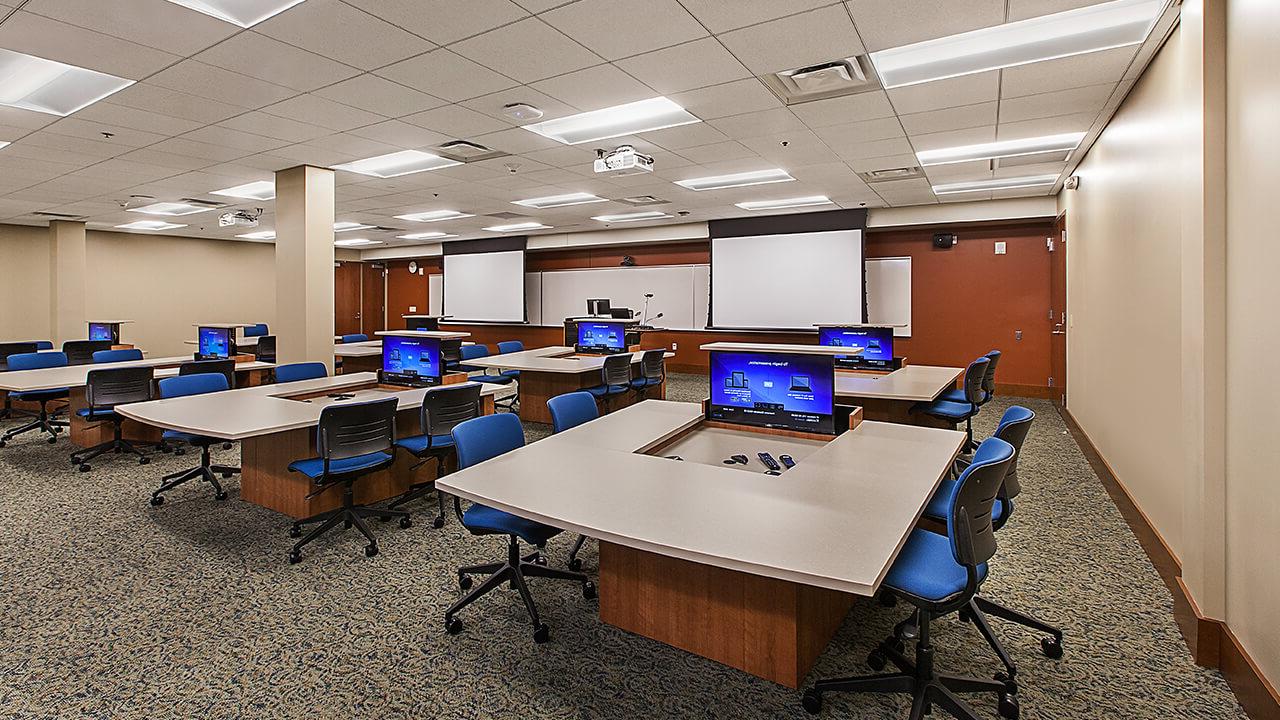 A large classroom with six breakout desks, each with a display protruding from the tabletop, and four chairs; two on each side. At the front of the classroom is a teaching station, flanked by projectors, and a white board almost spanning the width of the room.